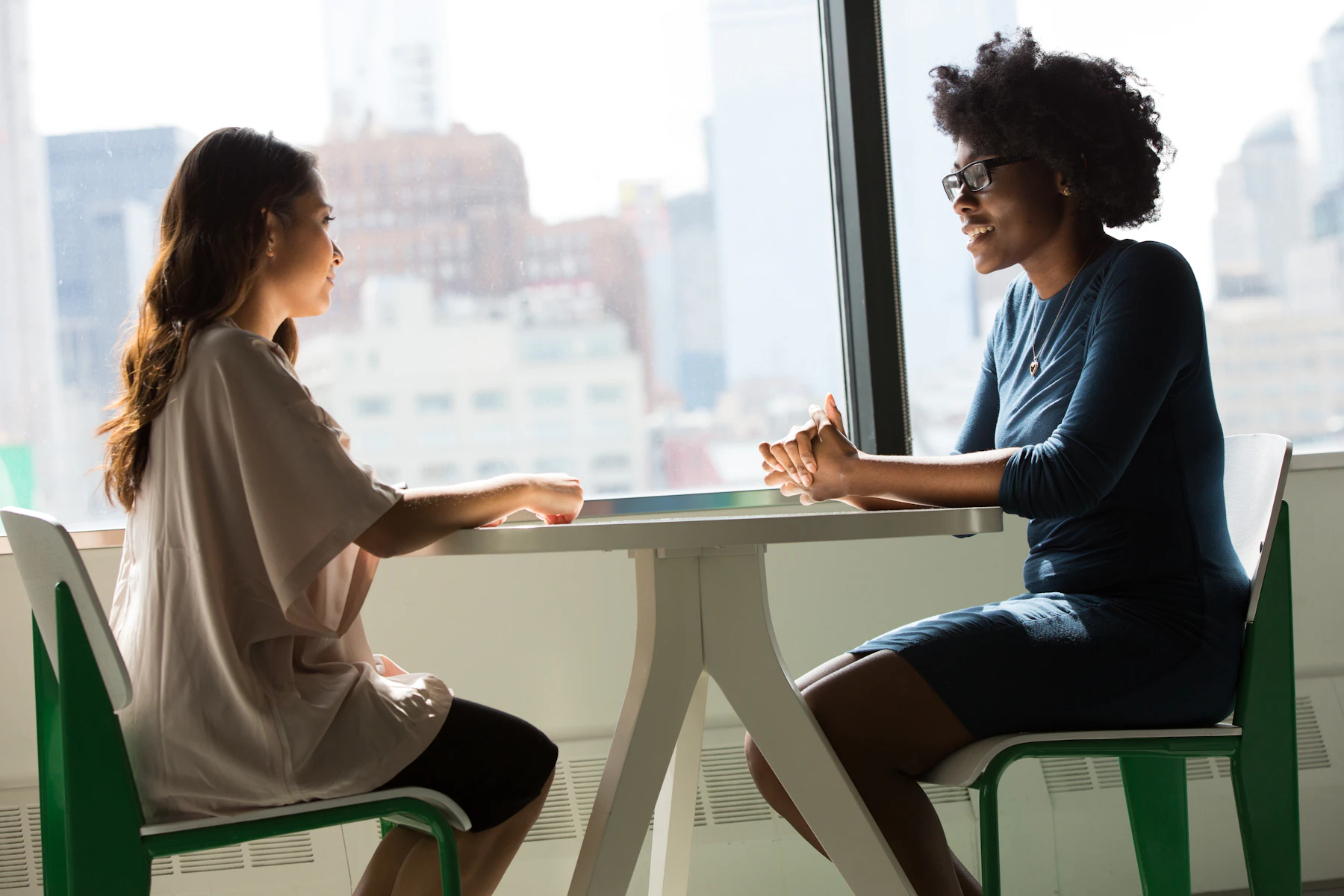 Two women having an attentive one-to-one conversation by a bright office window.